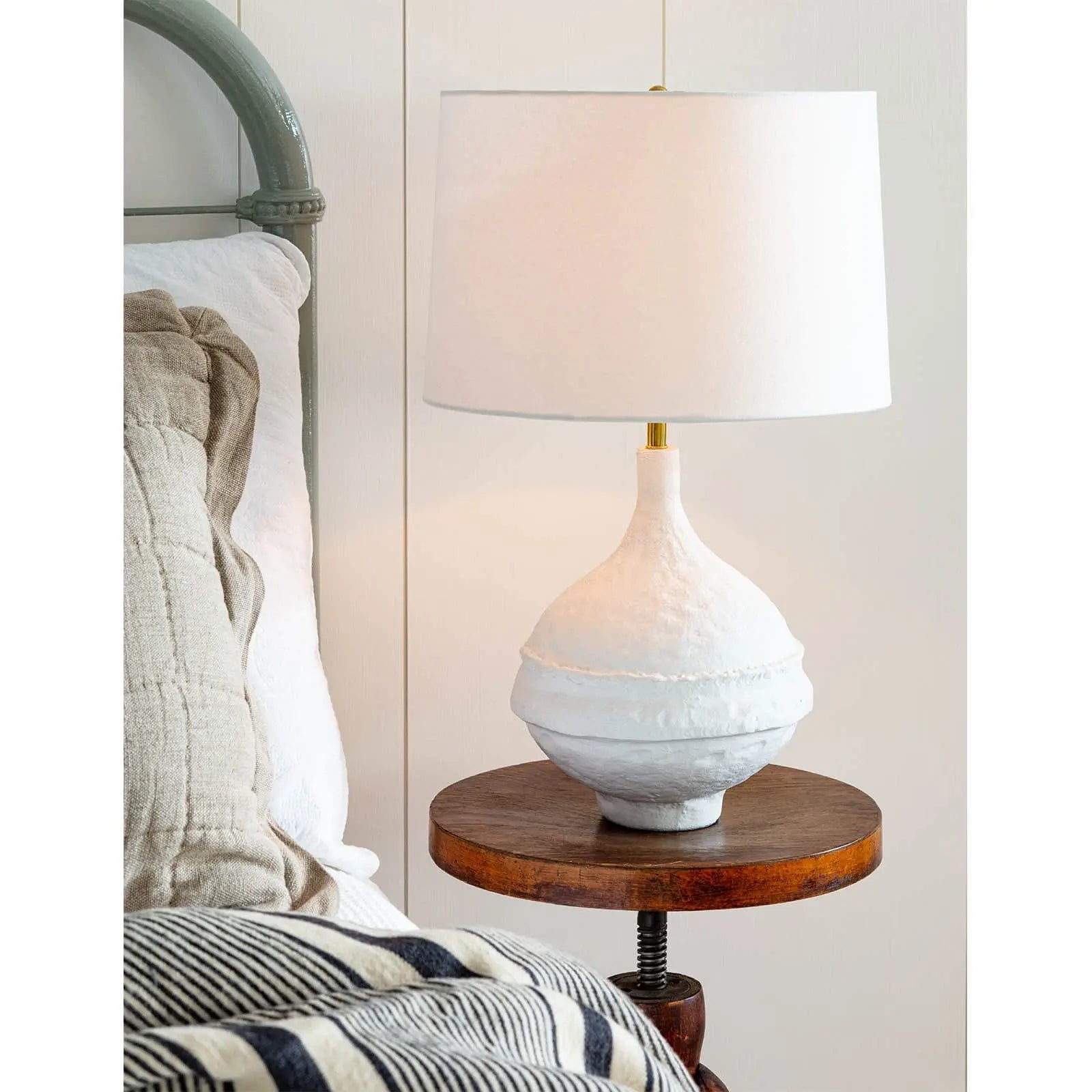 White ceramic lamp on a wooden side table next to a bed with striped bedding.