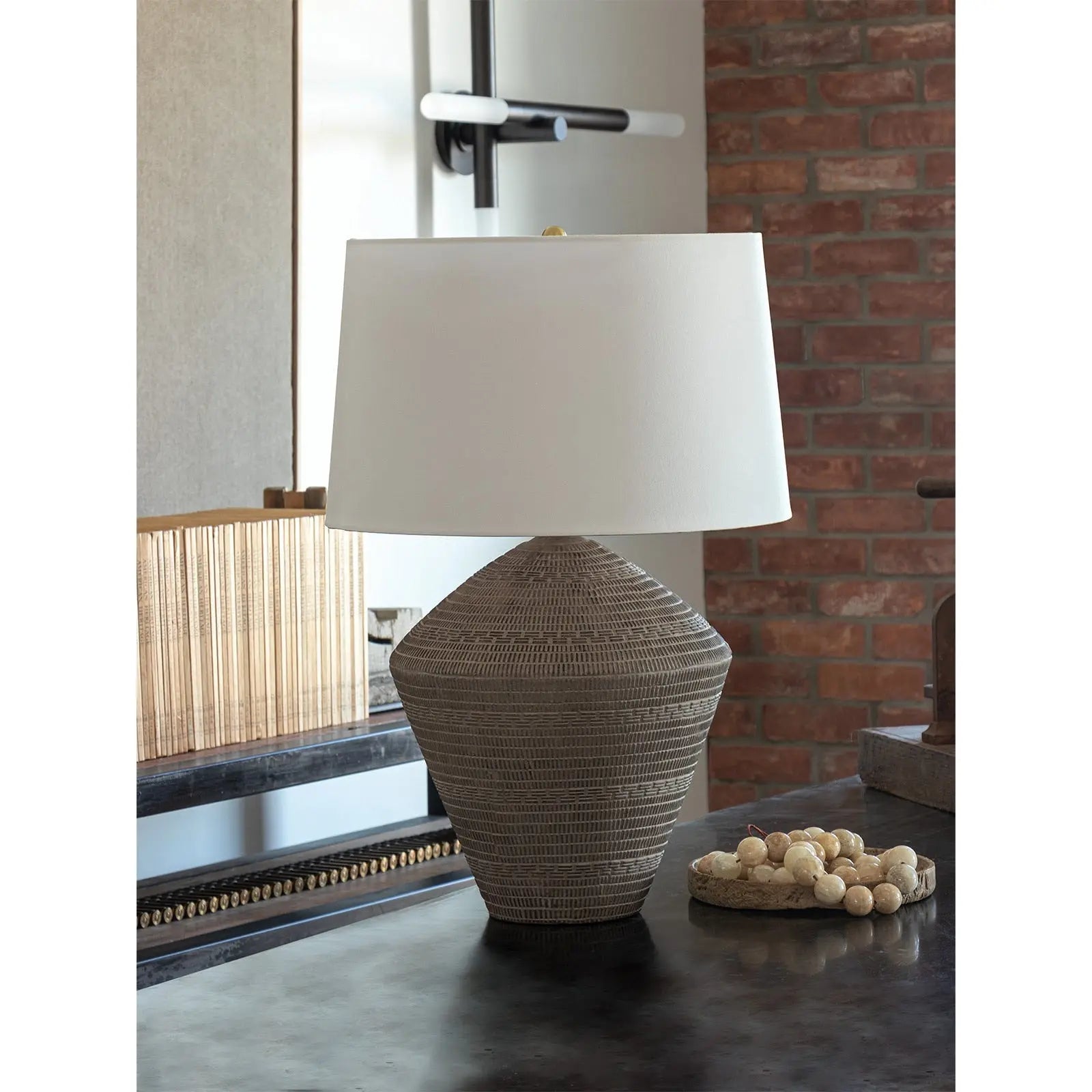 Table lamp with textured base and white shade on a surface with a brick wall background