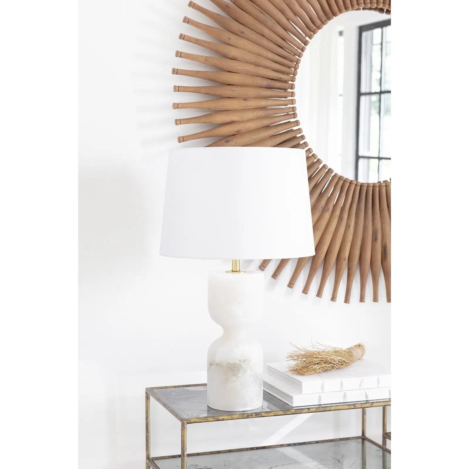 Decorative lamp with a white shade on a marble table, next to a sunburst mirror.
