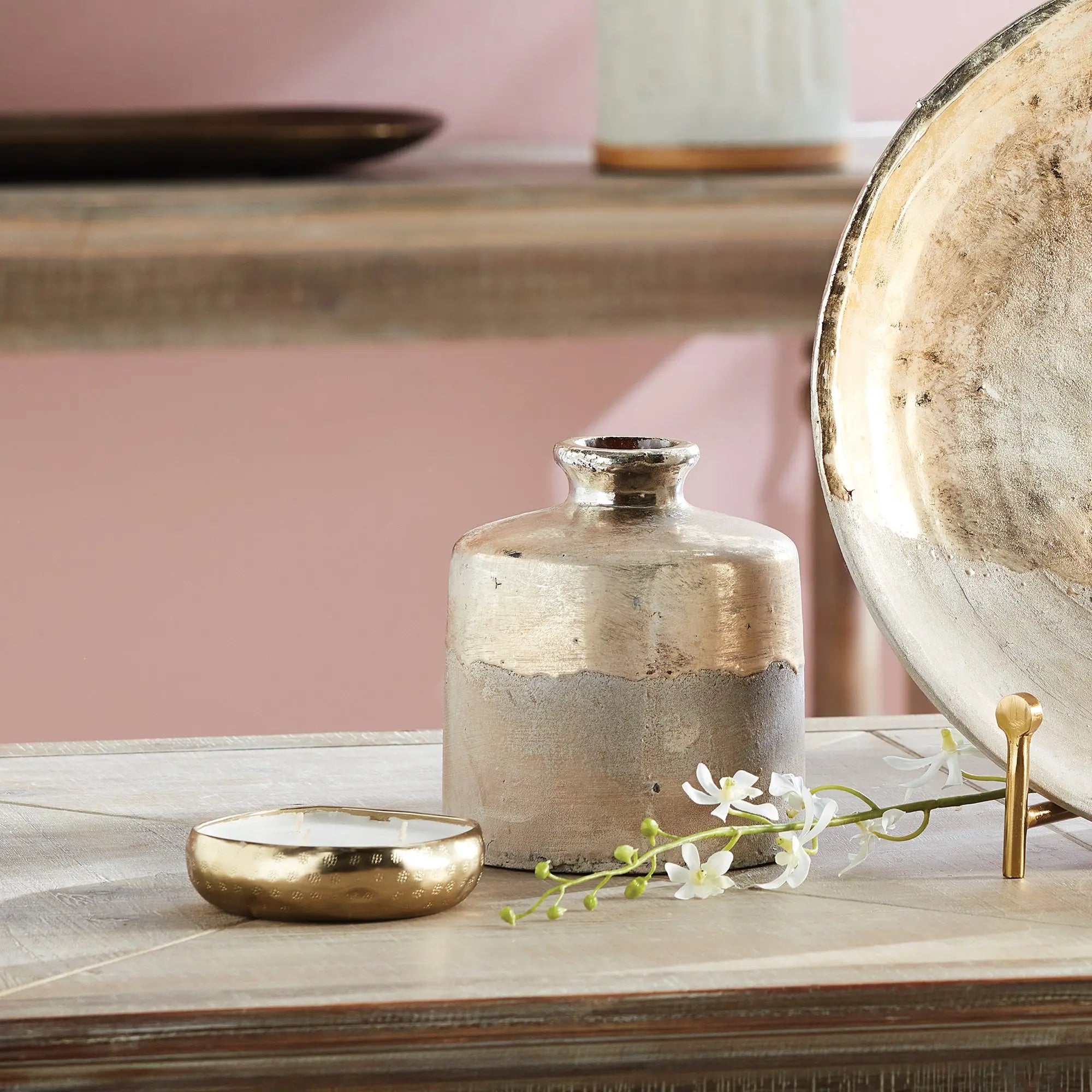 Decorative items including a small vase, gold dish, and floral arrangement on a wooden surface with a pink wall in the background.
