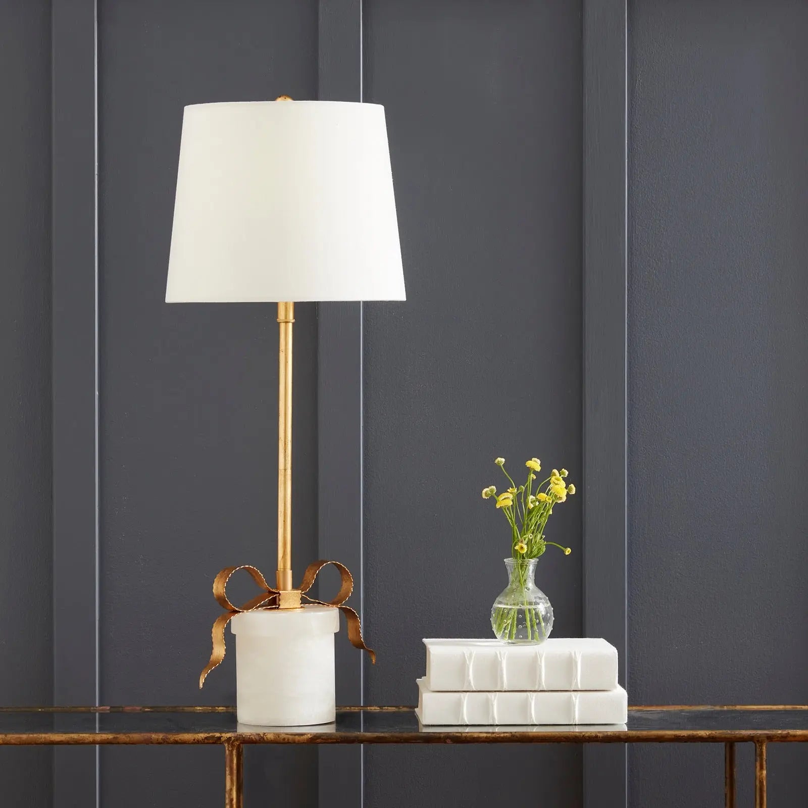 Decorative lamp with a white shade and gold base on a wooden surface against a dark gray wall.