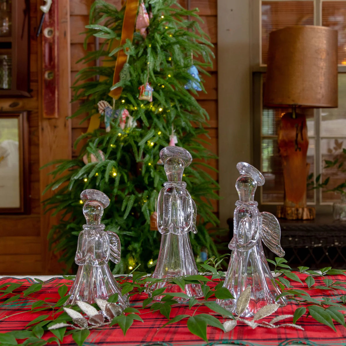 Three clear glass angel figurines with wings and hats are arranged on a red plaid tablecloth with green leaves, in front of a decorated Christmas tree and rustic indoor room background.